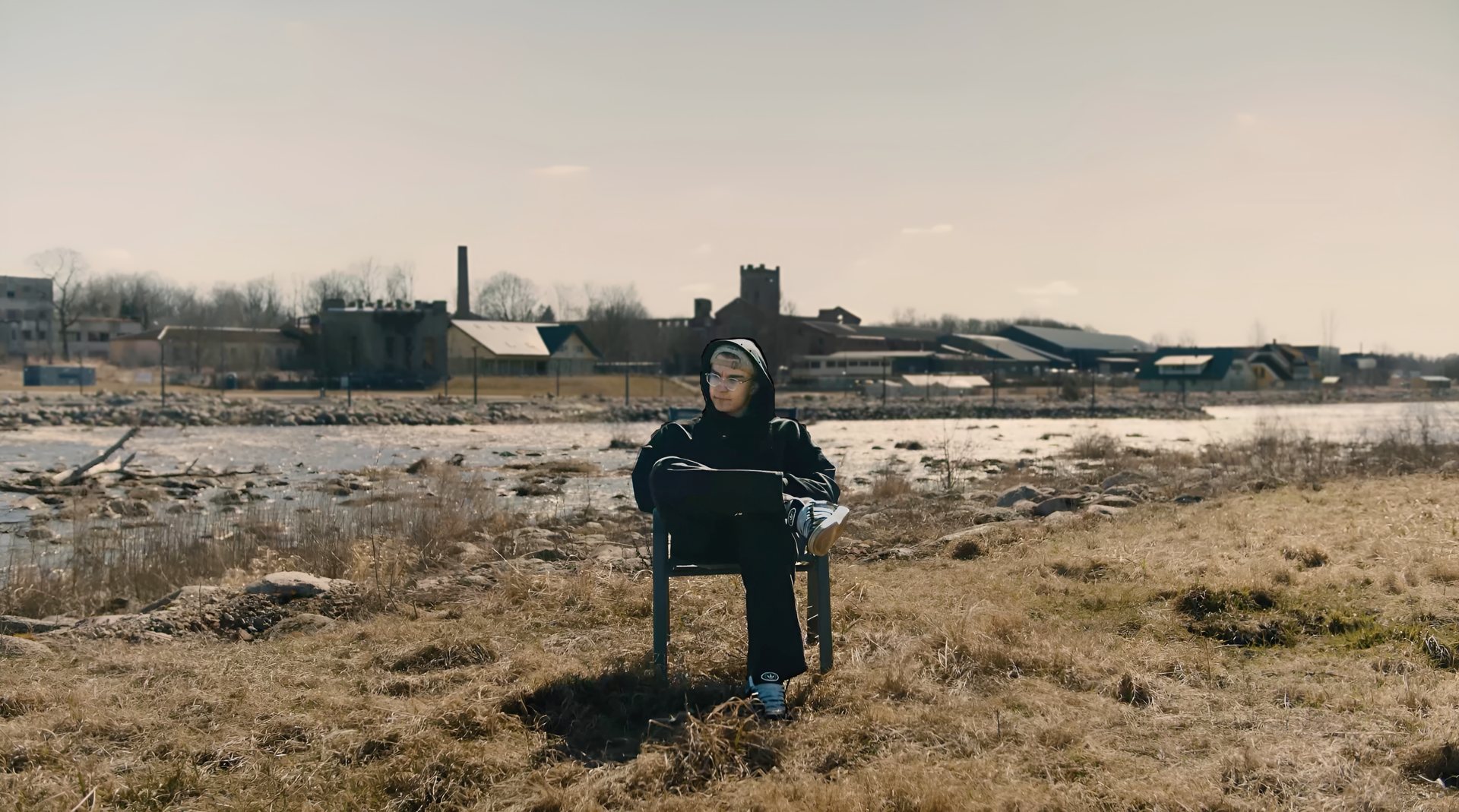 Andre Aadamsoo in a black hoodie, sitting on a chair in a dry field by a river, old factory buildings behind him